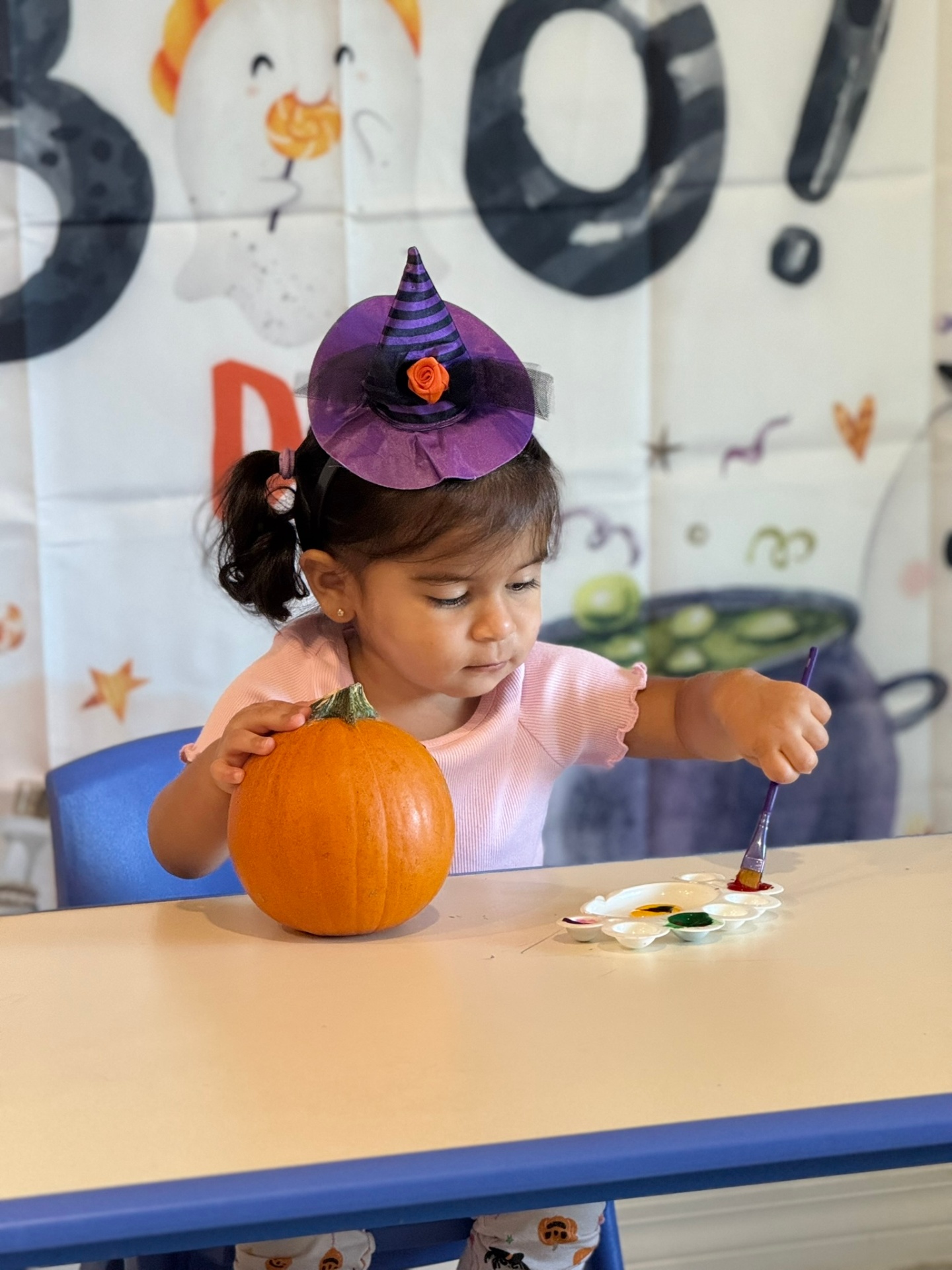 Toddler in a witch hat painting a pumpkin at a Halloween craft table