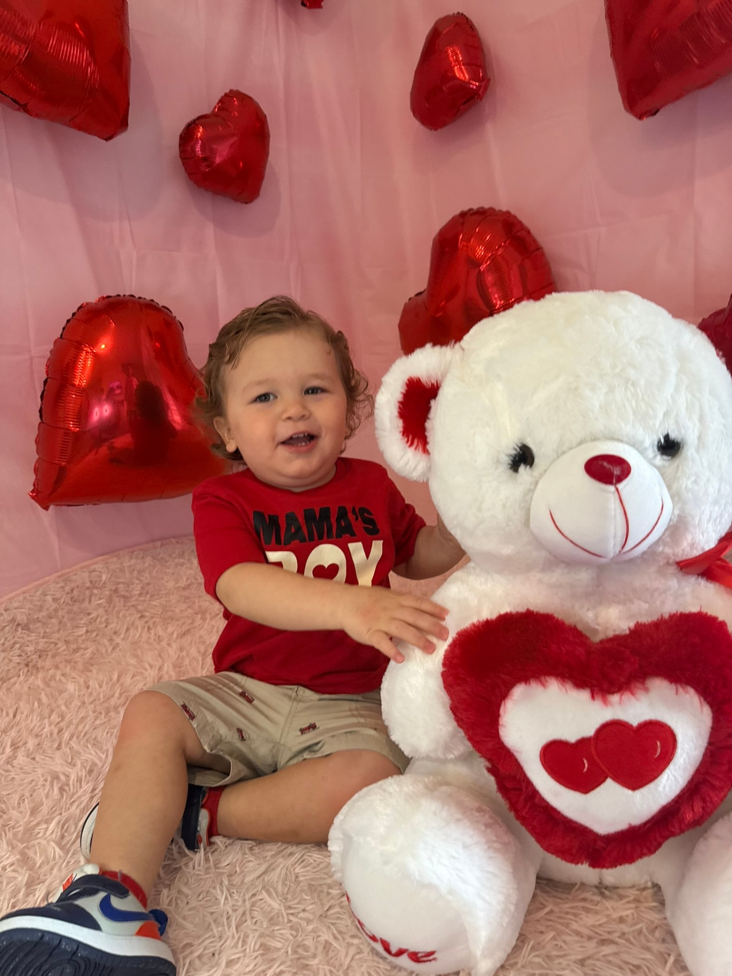 Toddler in a red Mama's Boy shirt sitting next to a large white teddy bear with red heart balloons