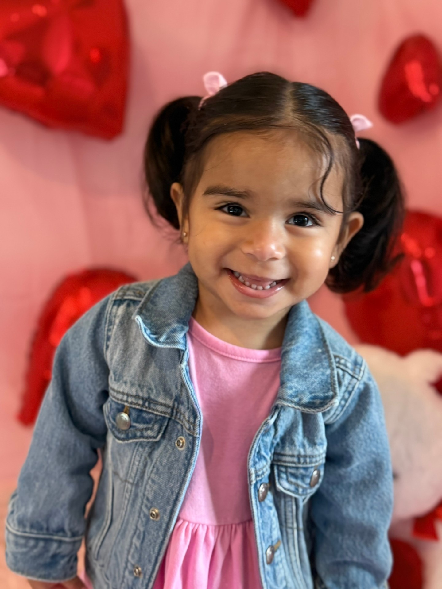 Smiling toddler with pigtails in a denim jacket in front of red heart balloons